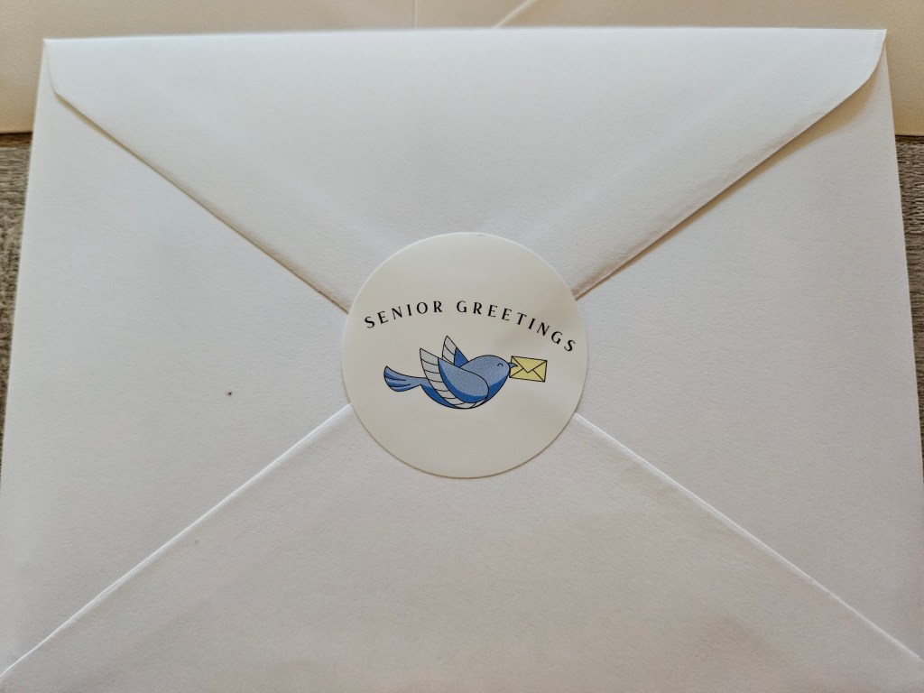 A white envelope with a round sticker label that says 'SENIOR GREETINGS' featuring a blue bird carrying an envelope.